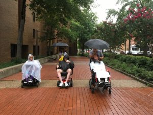 Image of wheelchair users with rain capes and umbrellas